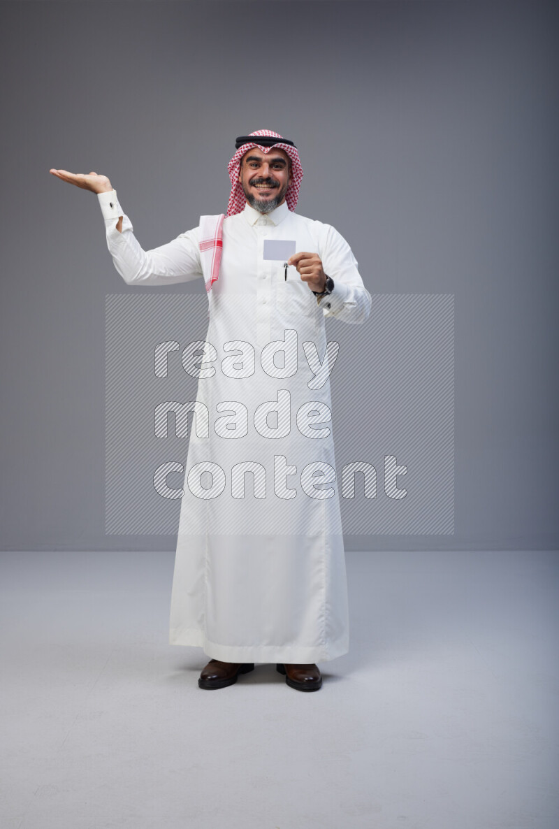 Saudi man Wearing Thob and red Shomag standing holding ATM card on Gray background