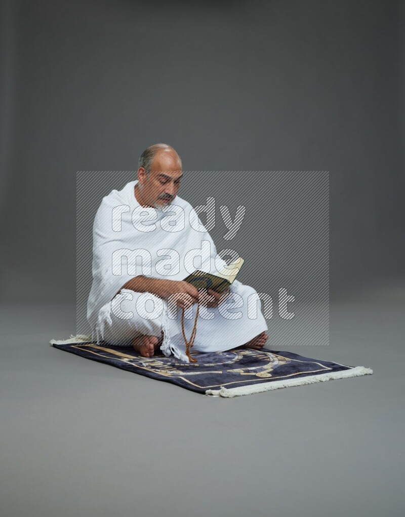 A man wearing Ehram sitting on mate prayer reading quran on gray background