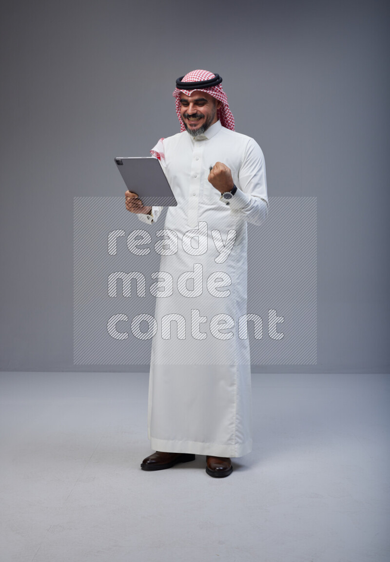 Saudi man Wearing Thob and red Shomag standing working on tablet on Gray background