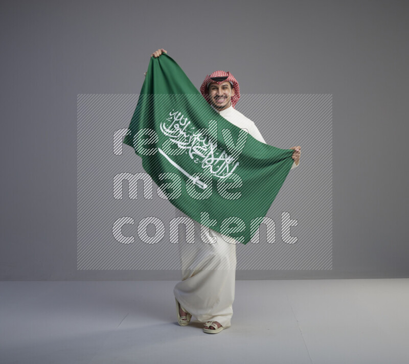 A saudi man standing wearing thob and red shomag holding big saudi flag on gray background