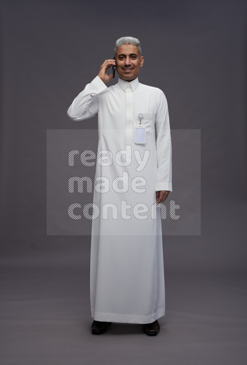 Saudi man wearing thob with pocket employee badge standing talking on phone on gray background