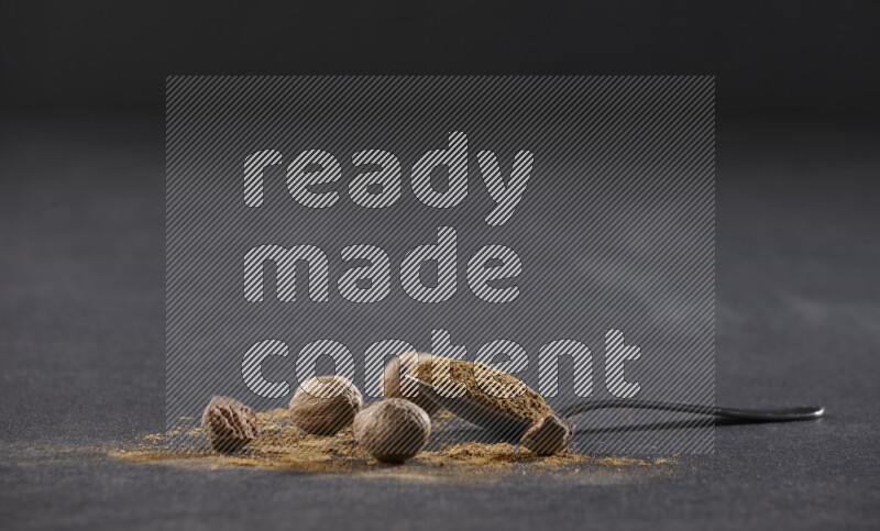 A metal spoon full of nutmeg powder with nutmeg powder and whole seeds beside it on a black flooring