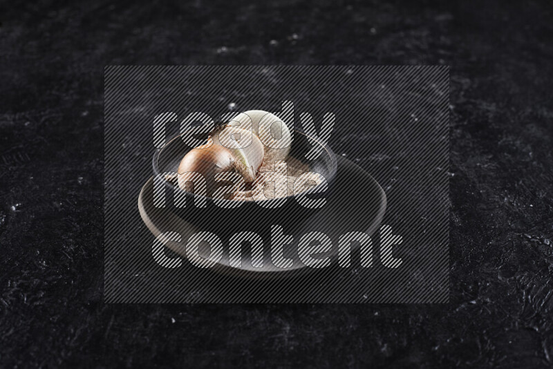 A black pottery bowl full of onion powder and some onion all on a pottery plate on black background