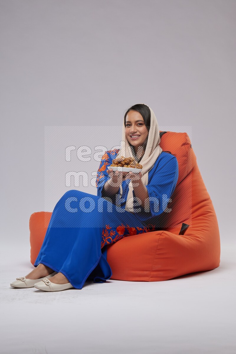 A Woman sitting on an orange beanbag wearing Jalabeya holding a plate of dates