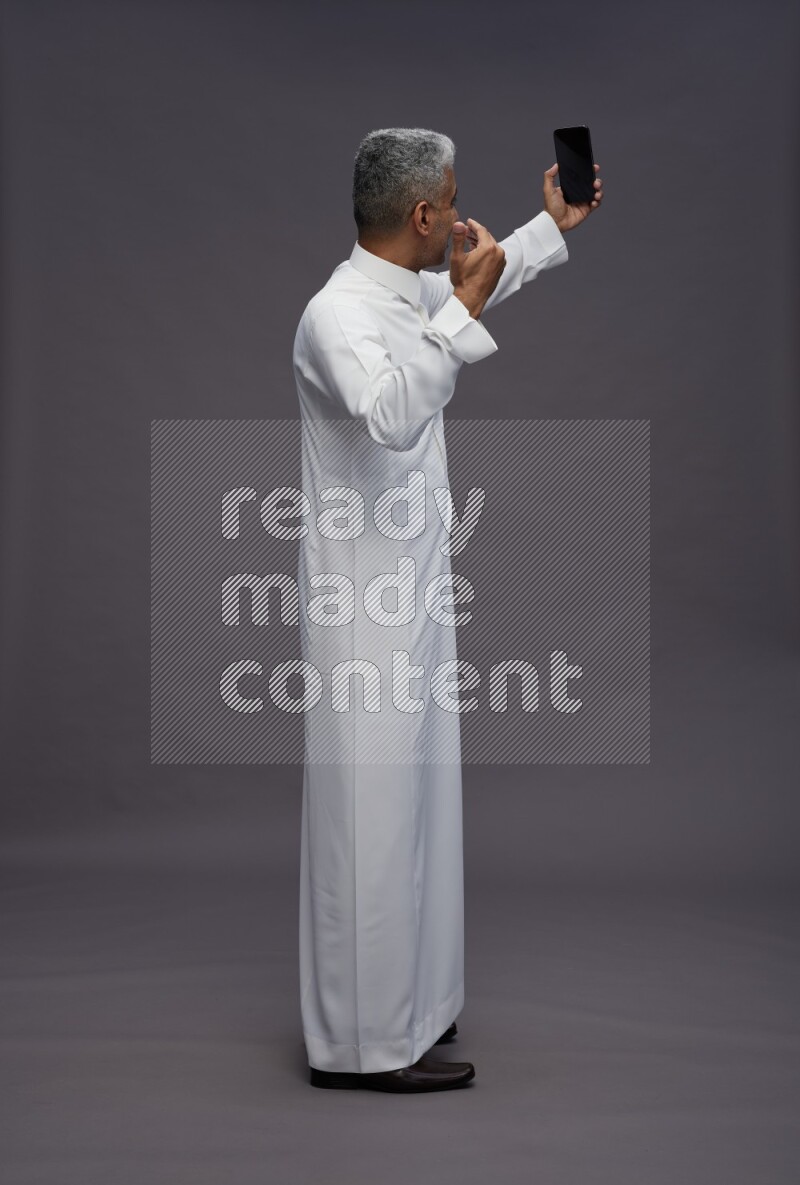 Saudi man wearing thob standing taking selfie on gray background