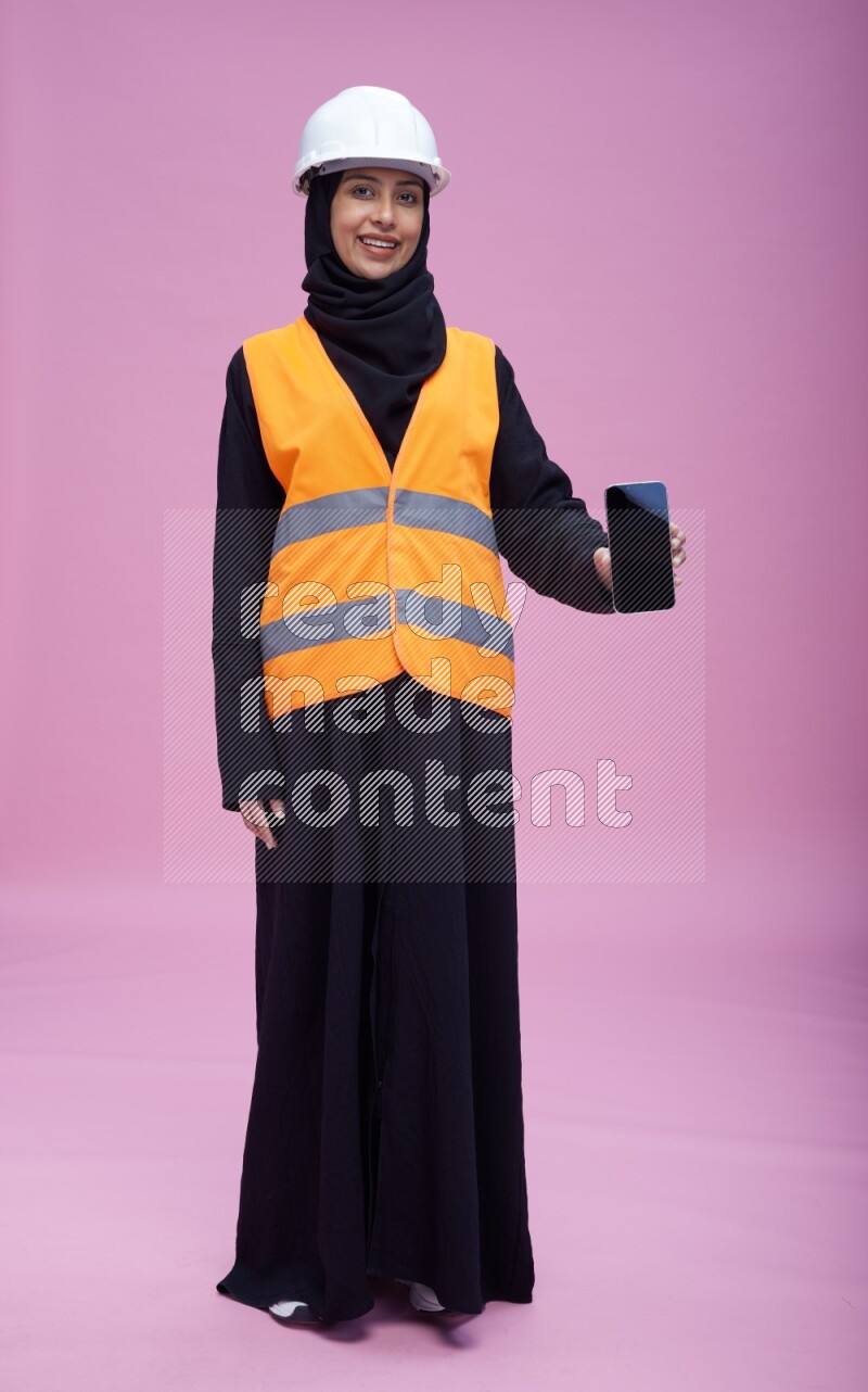 Saudi woman wearing Abaya with engineer vest and helmet standing showing phone to camera on pink background