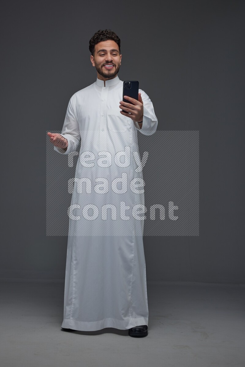 A Saudi man wearing Thobe and making a video call using his phone while standing and making different poses eye level on a gray background