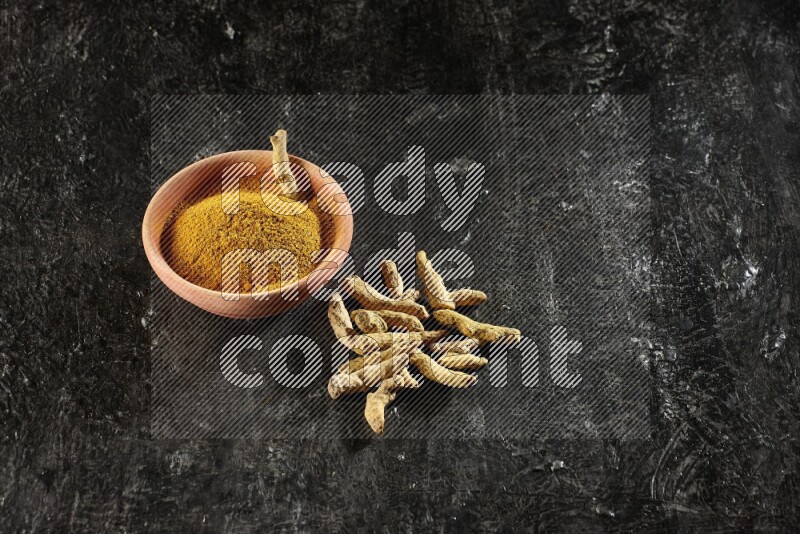 A wooden bowl full of turmeric powder with dried turmeric whole fingers on textured black flooring