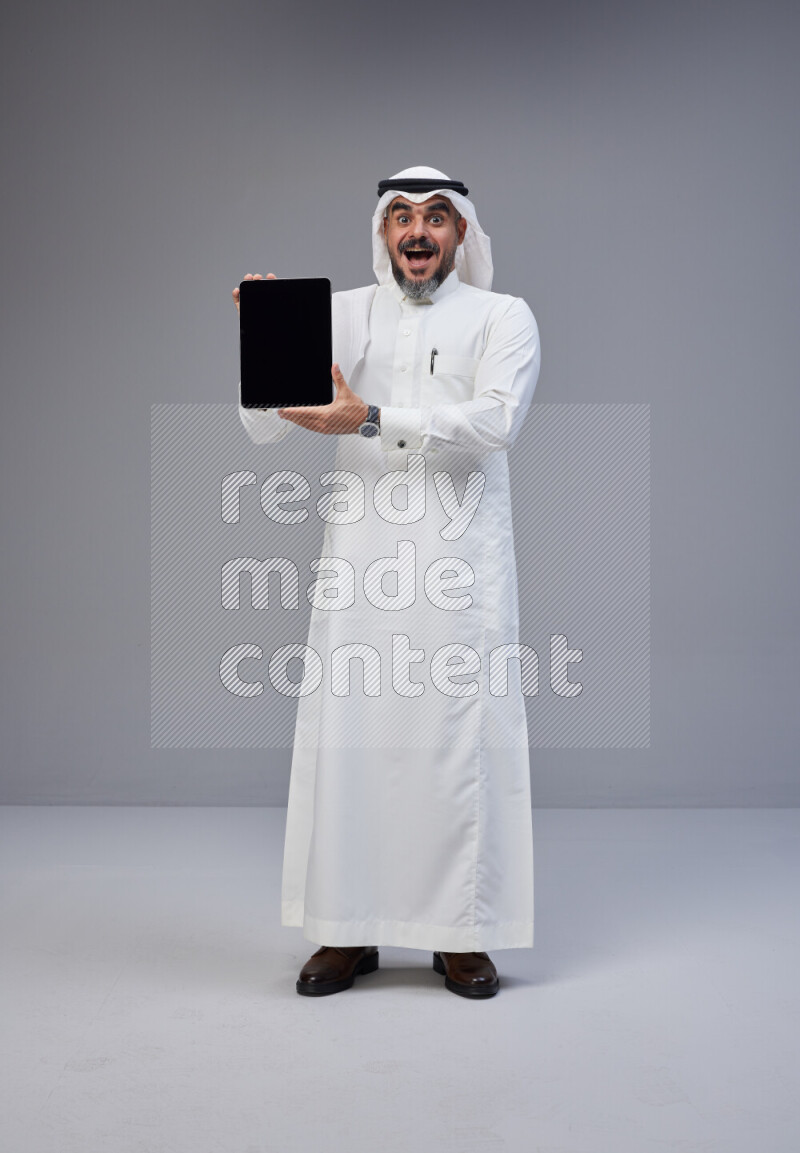 Saudi man Wearing Thob and white Shomag standing showing tablet to camera on Gray background