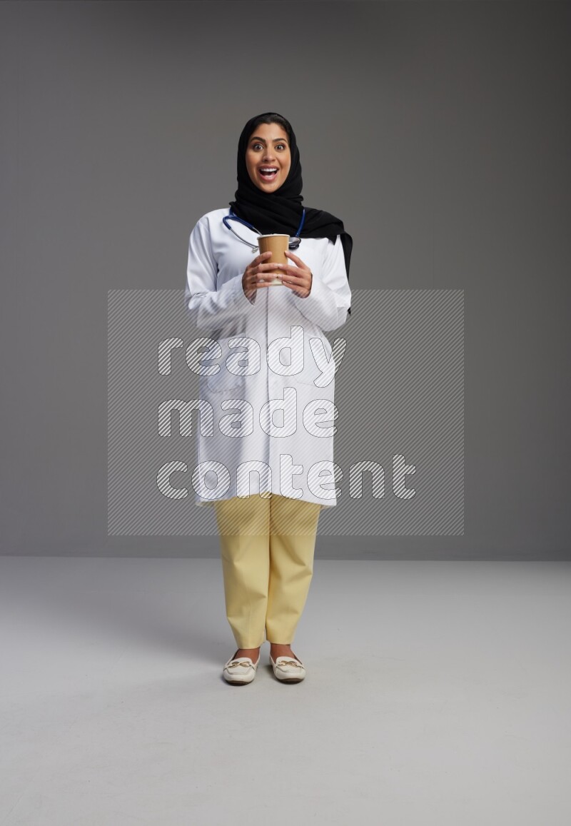 Saudi woman wearing lab coat with stethoscope standing holding paper cup on Gray background