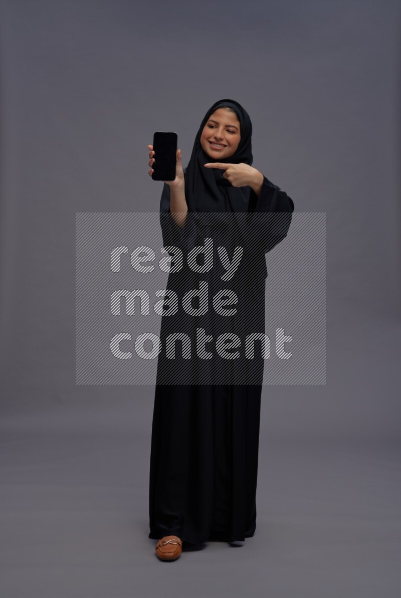 Saudi woman wearing Abaya standing showing phone to camera on gray background