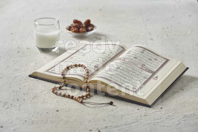 Quran with dates, prayer beads and different drinks all placed on textured white background