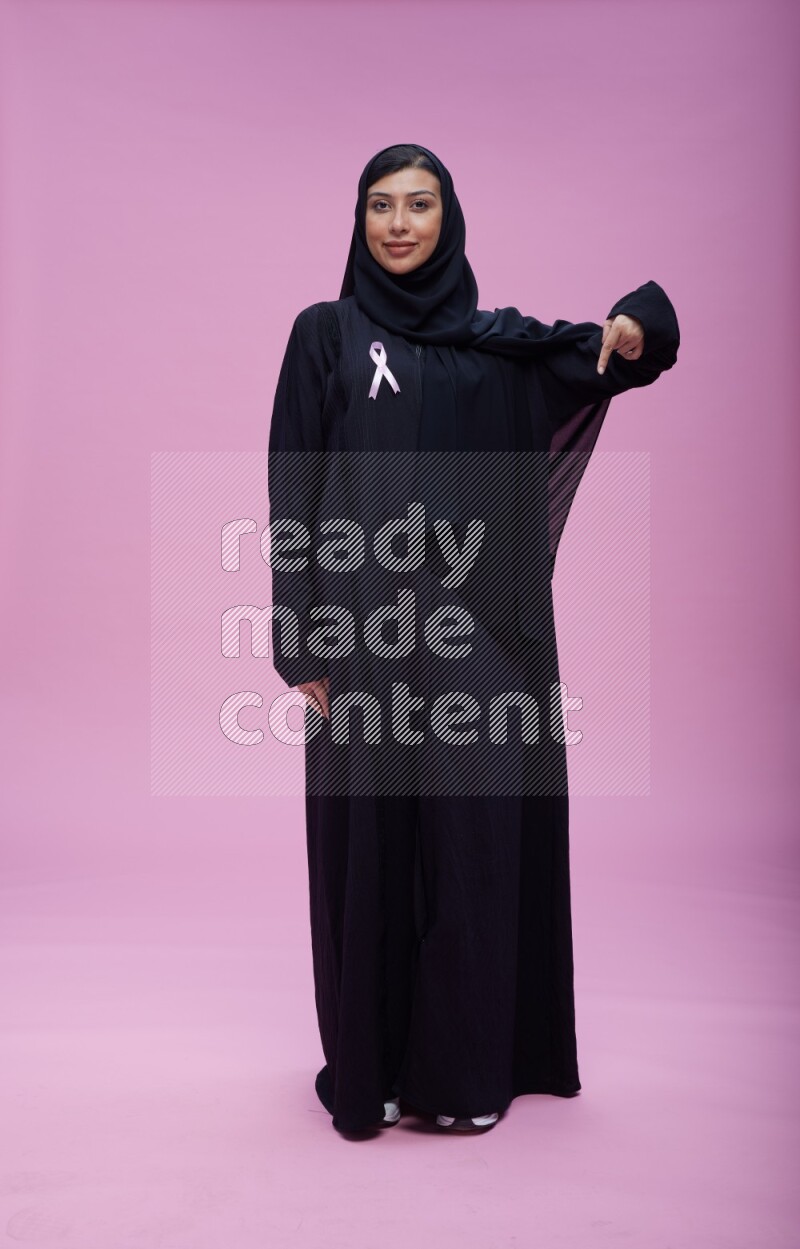 Saudi woman wearing pink ribbon on Abaya standing interacting with the camera on pink background