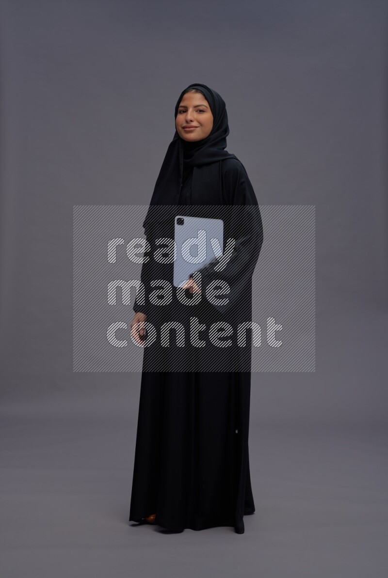Saudi woman wearing Abaya standing working on tablet on gray background