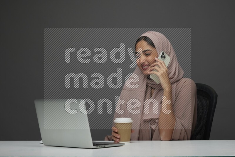 A Saudi woman Setting on her desk calling  on a Gray Background wearing Brown Abaya with Hijab