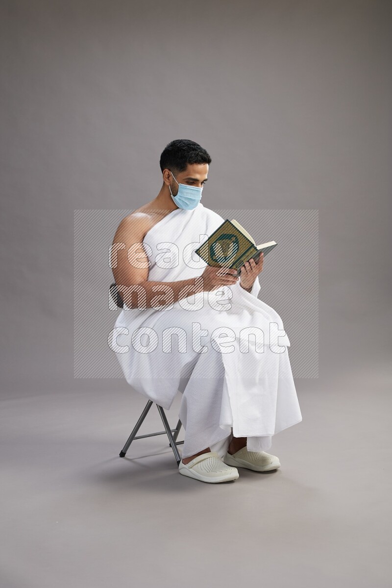 A man wearing Ehram with face mask sitting on chair reading quran on gray background