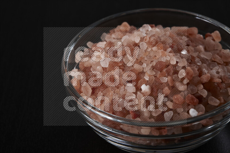 A glass bowl full of coarse himalayan salt crystals on black background