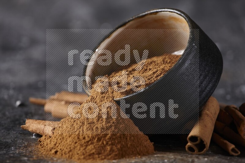 Black pottery bowl over filled with cinnamon powder and cinnamon sticks around the bowl on a textured black background