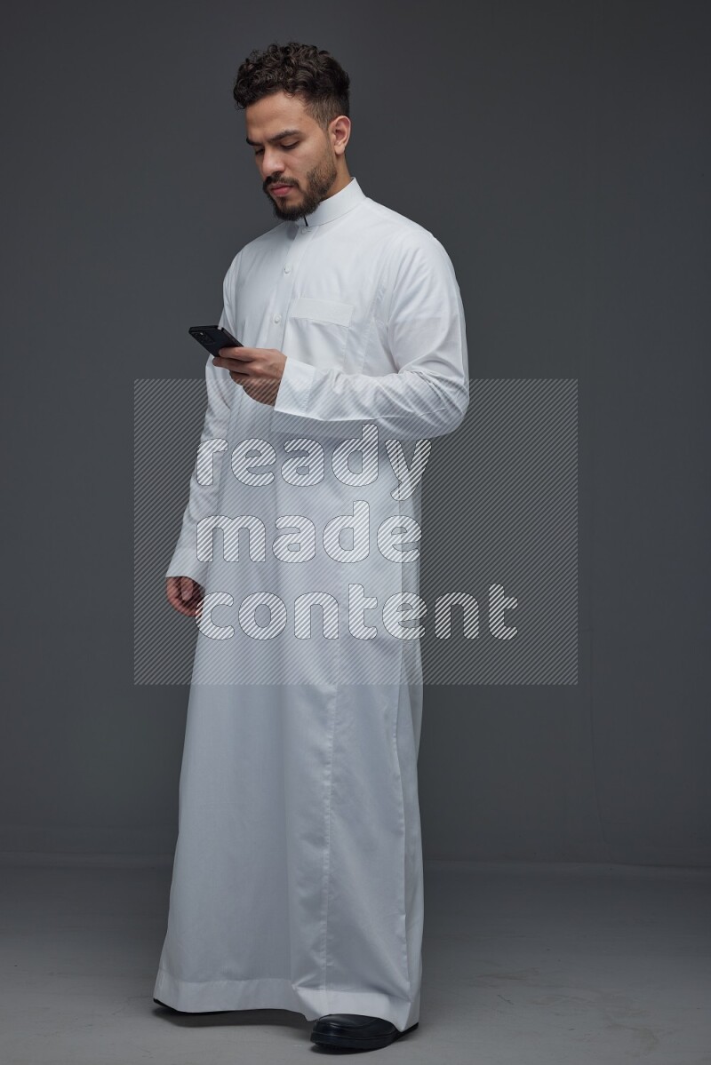 A Saudi man wearing Thobe standing and using his phone eye level on a gray background