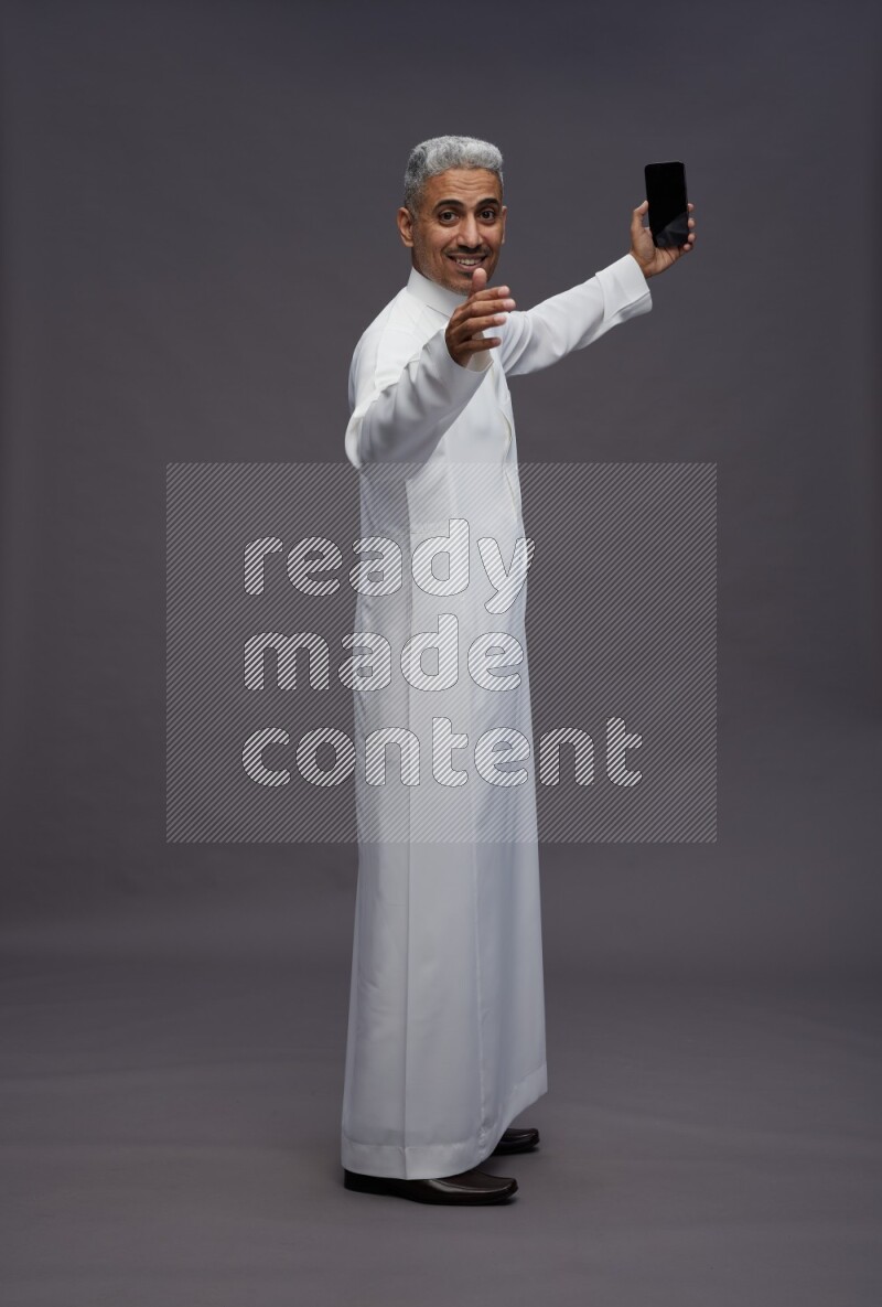 Saudi man wearing thob standing taking selfie on gray background
