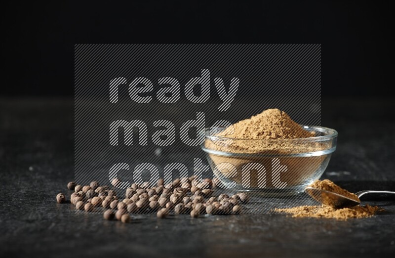 وعاء زجاجي وملعقة معدنية ممتلئان ببودرة بهار الفلفل الحلو وحبوبه متناثرة على خلفية سوداء