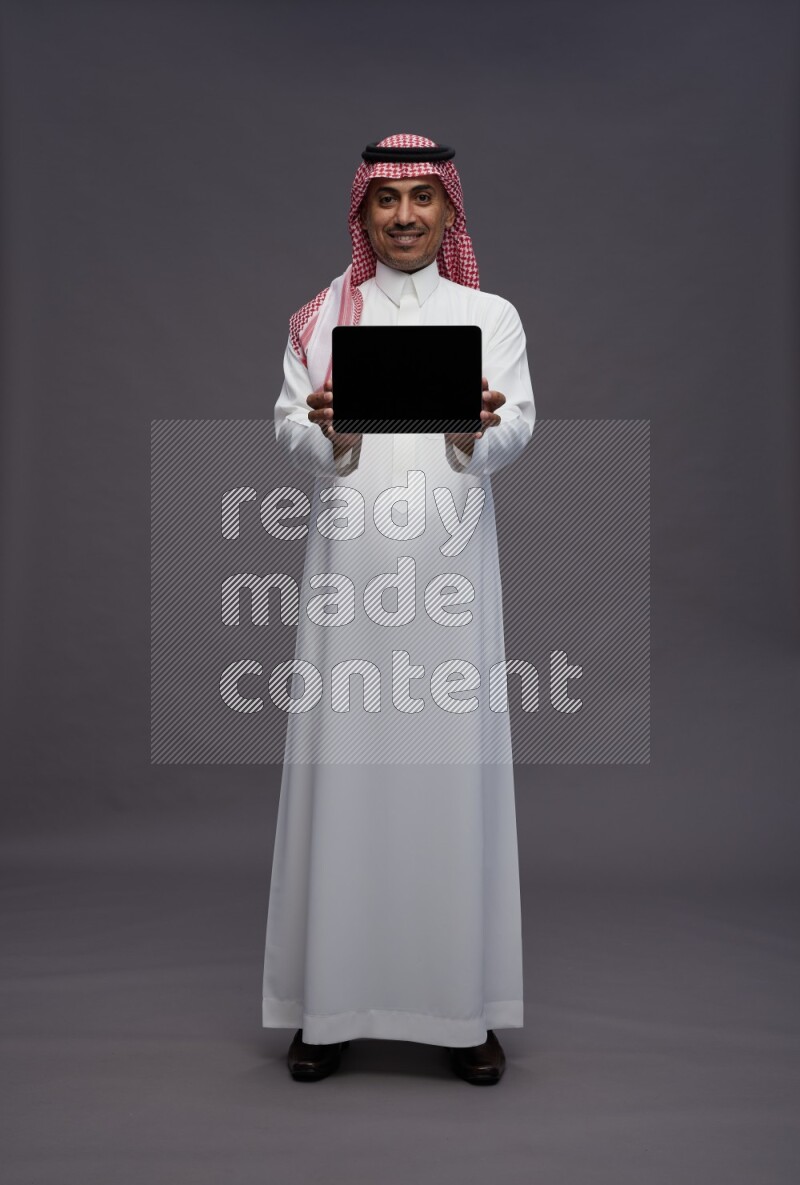 Saudi man wearing thob and shomag standing showing tablet to camera on gray background