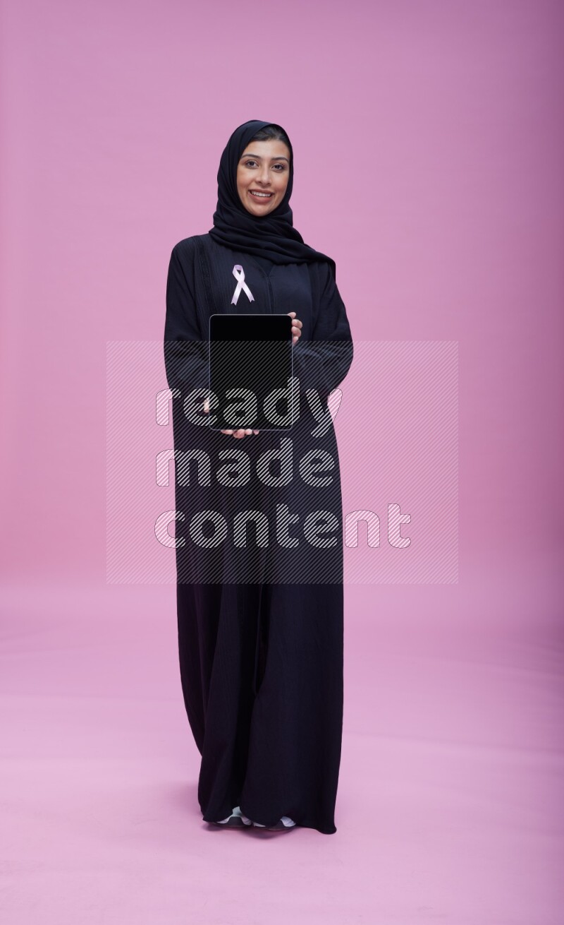 Saudi woman wearing pink ribbon on Abaya standing showing a tablet to camera on pink background