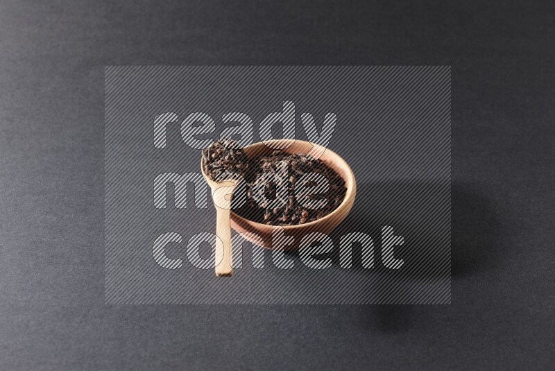 A wooden bowl and a wooden spoon full of cloves on a black flooring