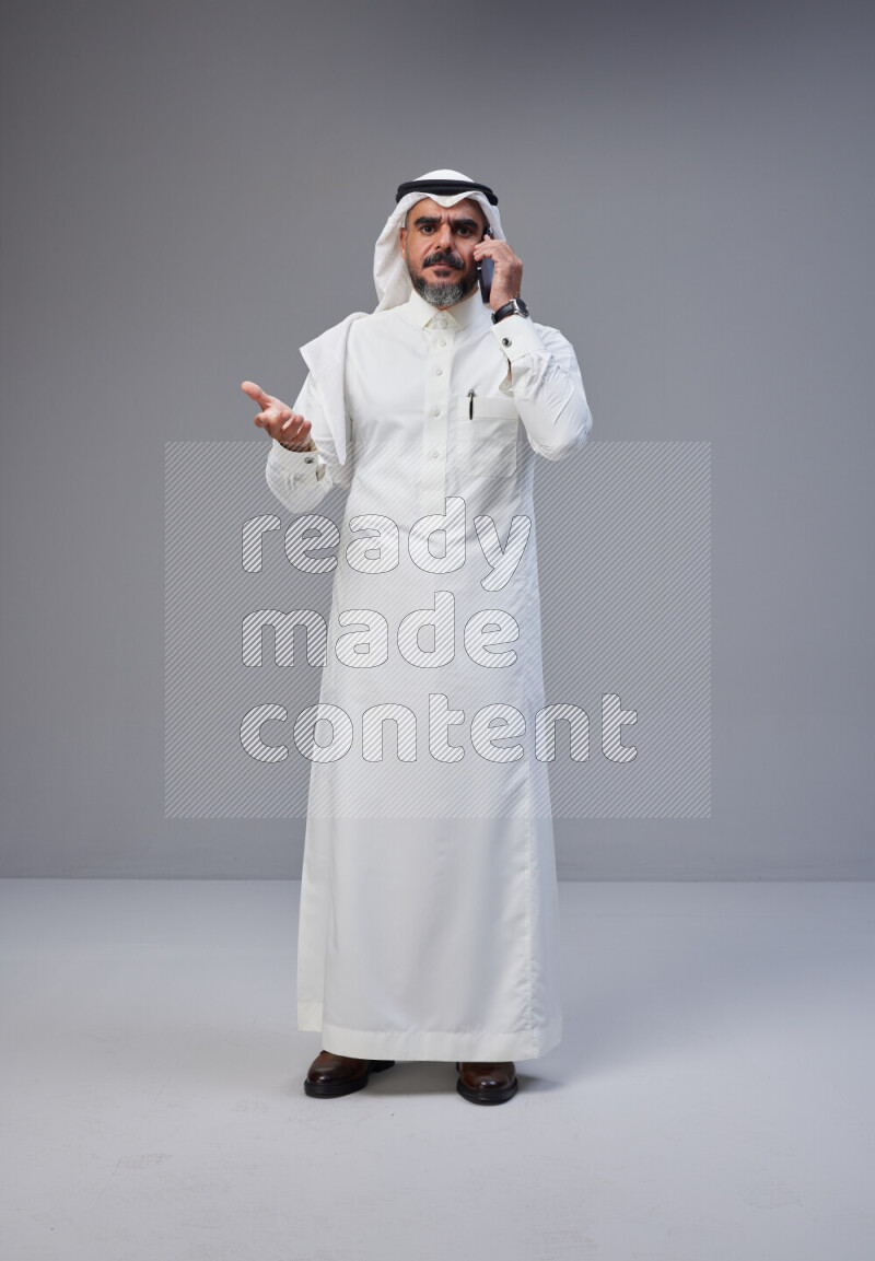 Saudi man Wearing Thob and white Shomag standing talking on phone on Gray background