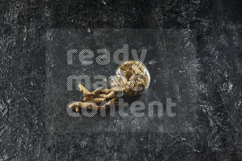 A flipped glass spice jar full of dried turmeric whole fingers and fingers fell out of it on textured black flooring