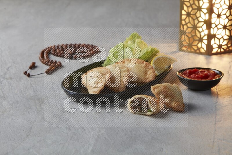 Three fried sambosas in an oval shaped black plate and a red sauce in a black round ramekin with a brown misbaha and a golden lantern on the side on a gray background