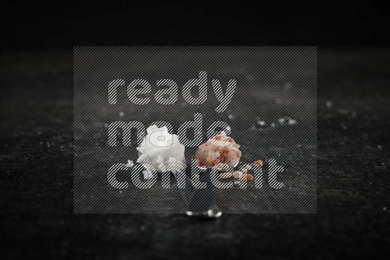 2 metal spoons filled with white salt and pink himalayan salt on black background