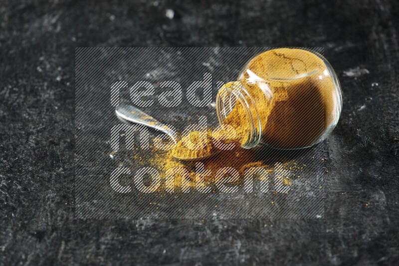 وعاء زجاجي للتوابل مقلوب وملعقة معدنية ممتلئان ببودرة الكركم والبودرة تناثرت منه على خلفية سوداء