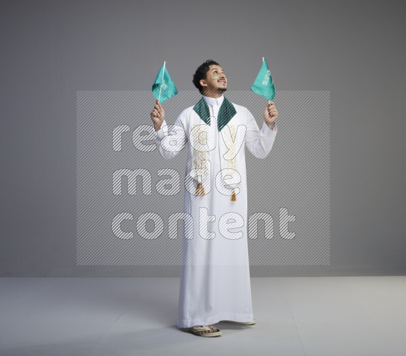 A Saudi man standing wearing thob and saudi flag scarf with face painting holding small Saudi flag on gray background
