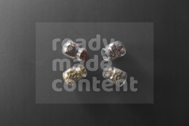 4 glass spice jars full of cloves, cardamom, basil and star anise on black flooring