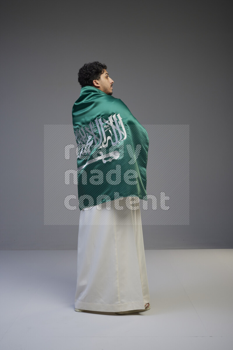 A Saudi man standing wearing thob wrapping big Saudi flag on gray background