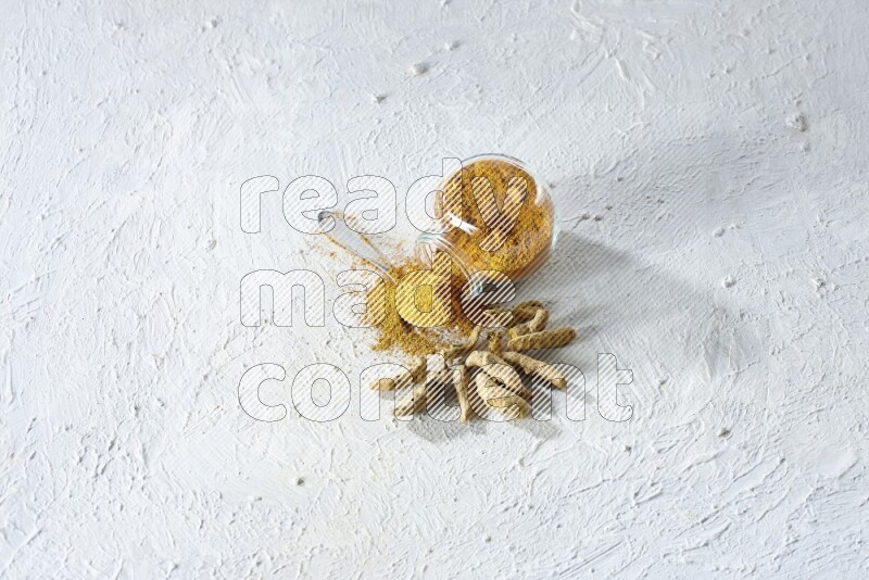 وعاء زجاجي للتوابل مقلوب وملعقة معدنية ممتلئة ببودرة الكركم والبودرة تناثرت منه مع أصابع الكركم المجففة على خلفية بيضاء