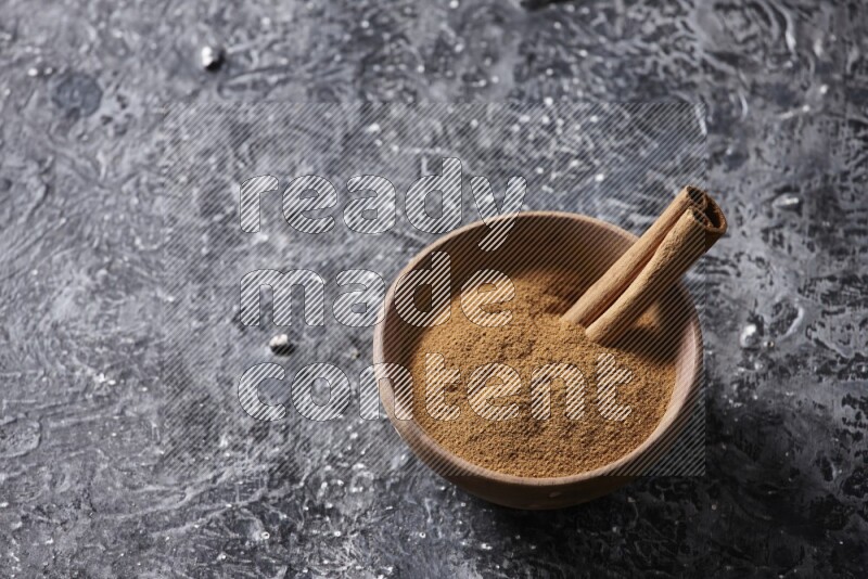 Wooden bowl full of cinnamon powder and a cinnamon stick on a textured black background