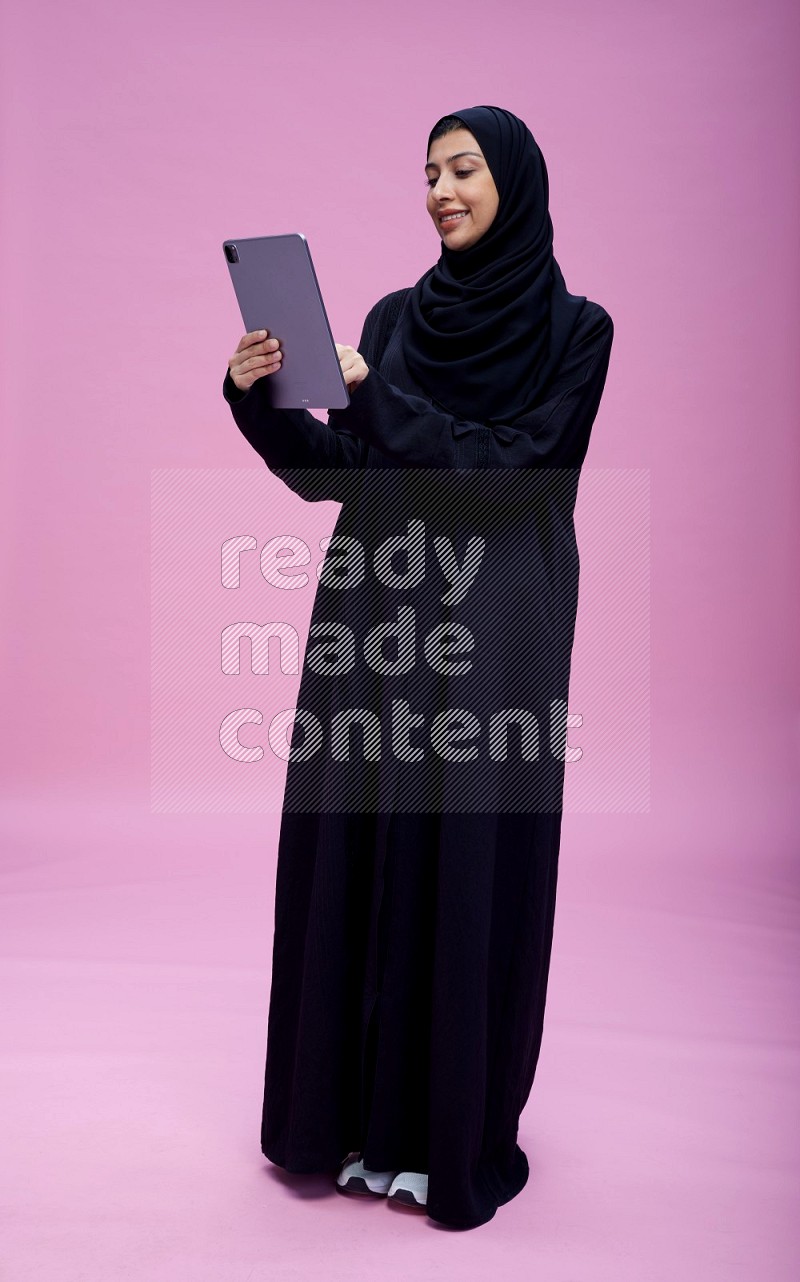 Saudi woman wearing Abaya standing working on tablet on pink background