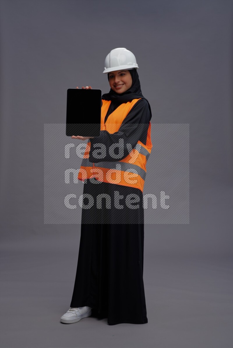 Saudi woman wearing Abaya with engineer vest standing showing tablet to camera on gray background
