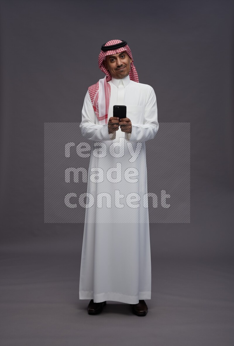 Saudi man wearing thob and shomag standing texting on phone on gray background