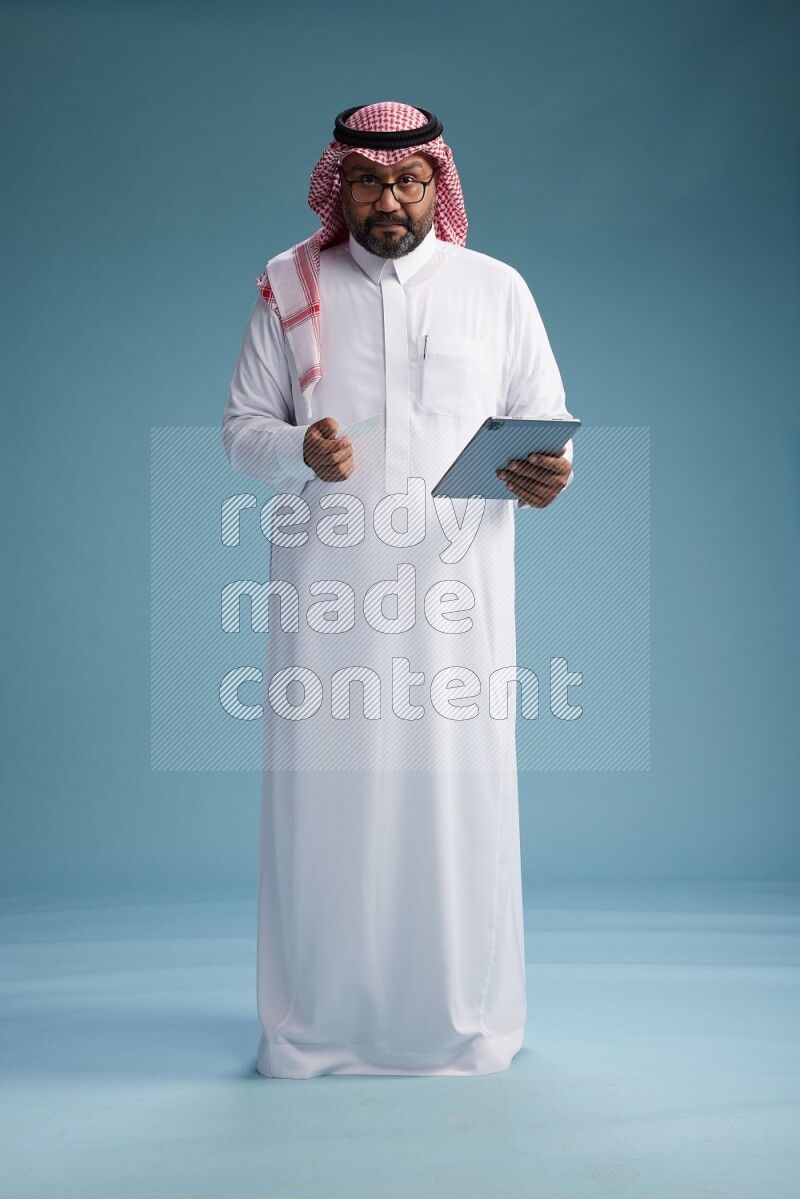 Saudi Man with shimag Standing holding ATM card while working on tablet on blue background