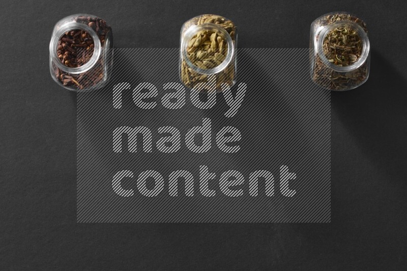 3 glass spice jars full of cloves, cardamom and basil on black flooring