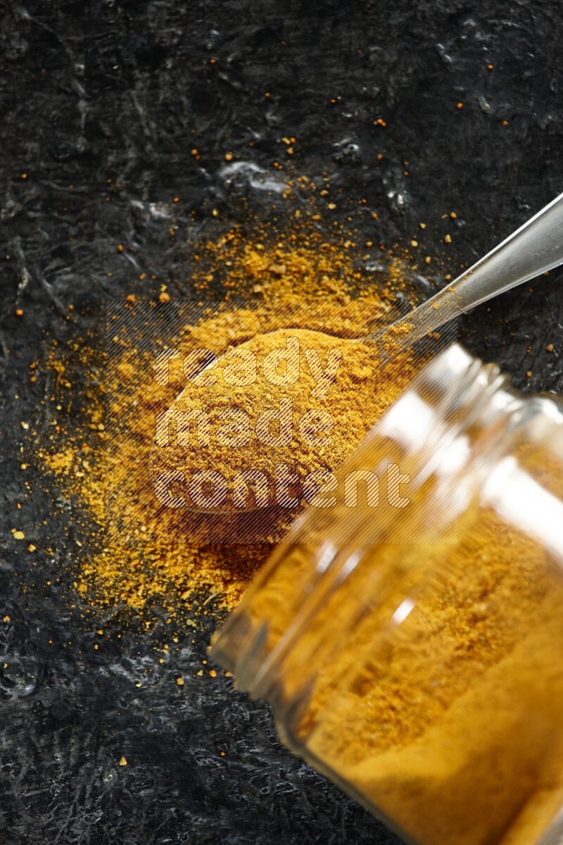 A flipped glass jar and a metal spoon full of turmeric powder and powder spilled out of it on textured black flooring