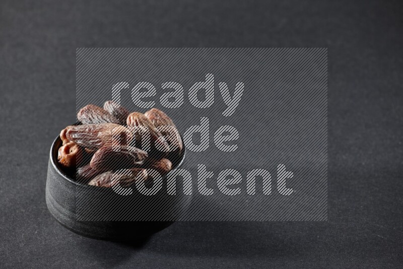 A black pottery bowl full of dried dates on a black background in different angles