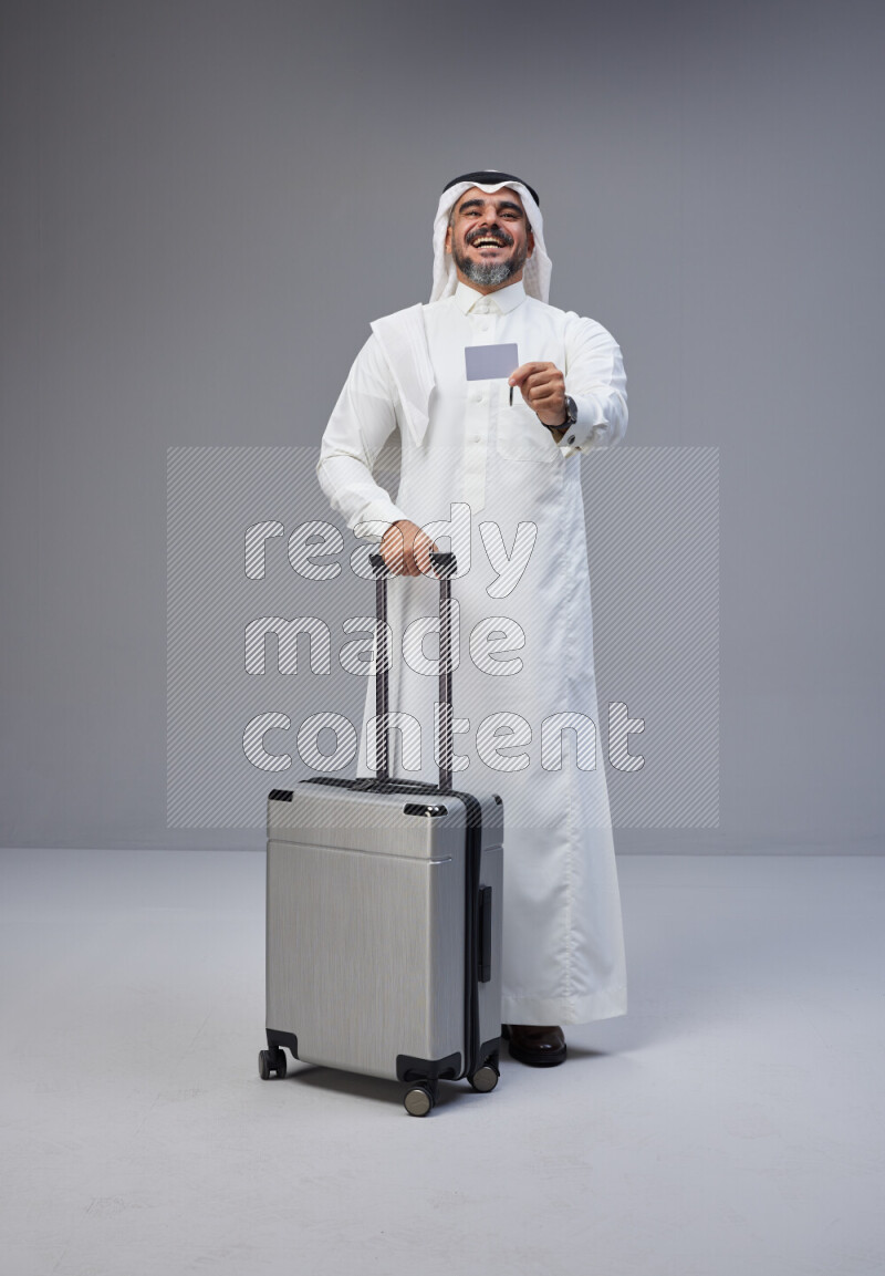 Saudi man wearing Thob and white Shomag standing holding Travel bag and ATM card on Gray background