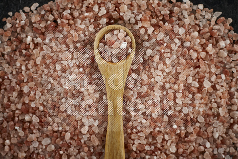 A wooden spoon full of coarse himalayan salt crystals on a bunch of the crystals on black background