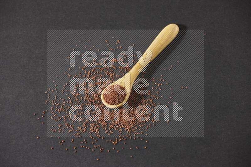 A wooden spoon full of garden cress seeds and seeds spread beside it on a black flooring