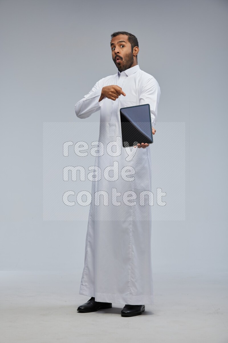 Saudi man wearing Thob standing showing tablet to camera on Gray background