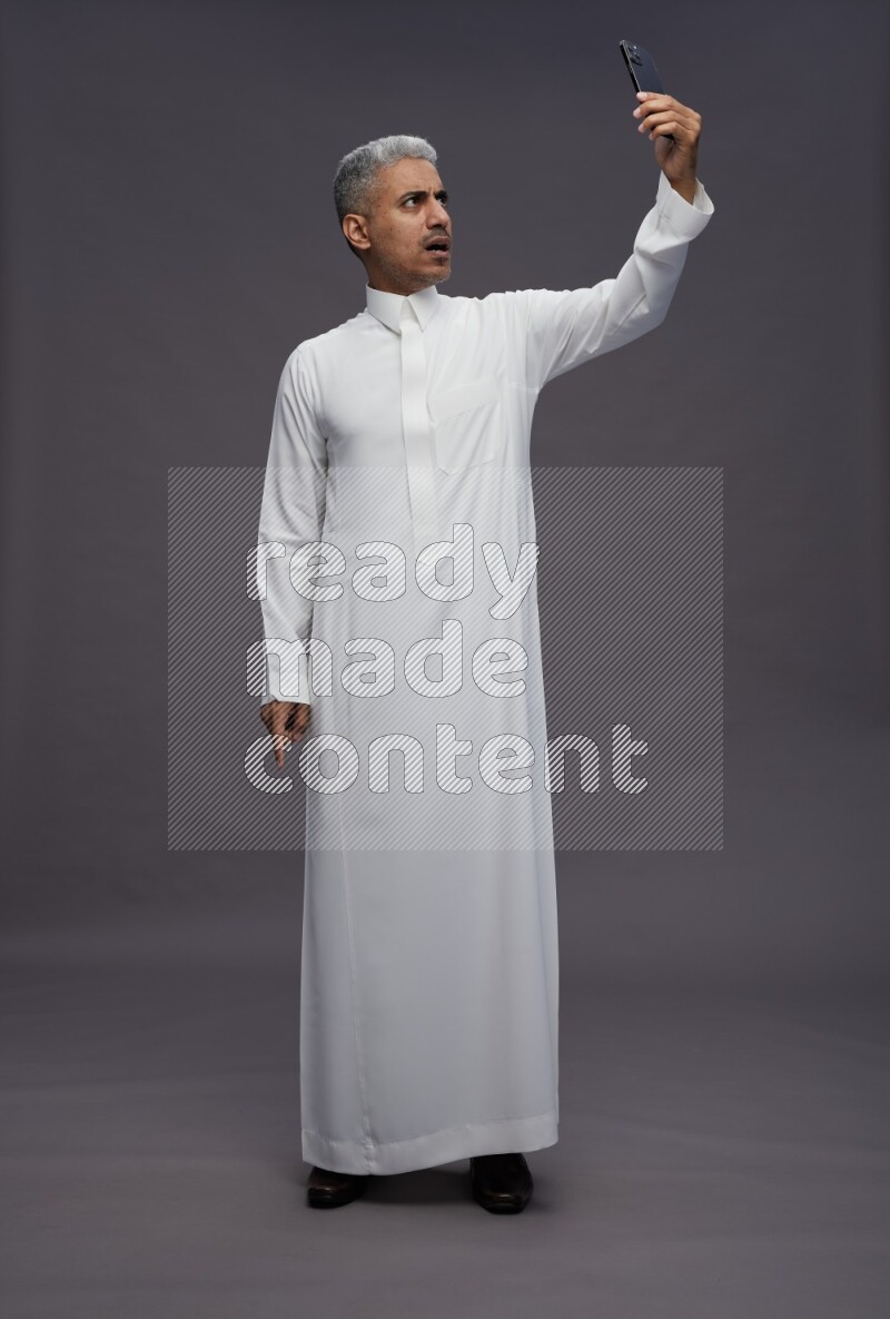 Saudi man wearing thob standing taking selfie on gray background